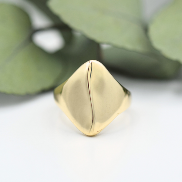 A gold signet ring with a textured, dune-shaped design, placed on a white surface next to green leaves.