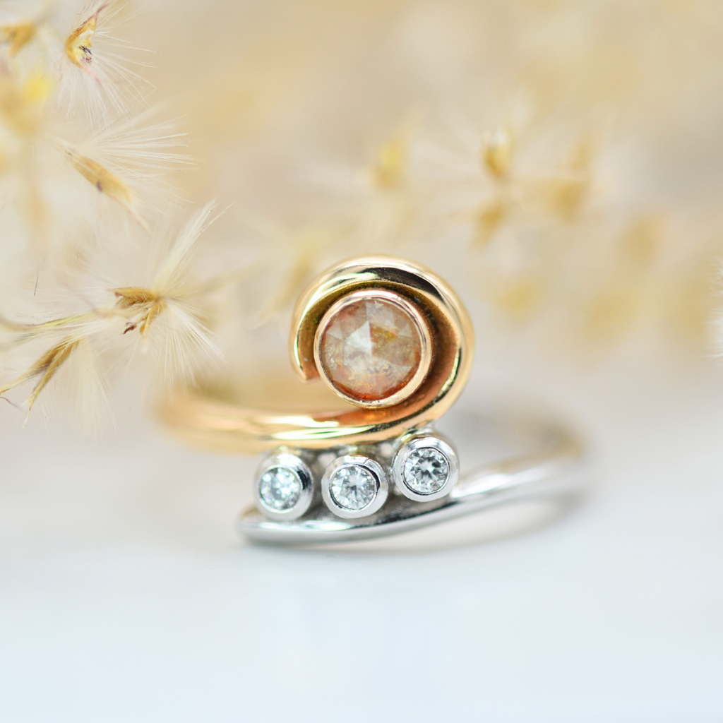 A rose gold ring with a round peach diamond centerpiece and three smaller round brilliant cut diamonds set on its side, photographed on a white background with dried flowers in the foreground.