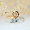 A rose gold ring with a round peach diamond centerpiece and three smaller round brilliant cut diamonds set on its side, photographed on a white background with dried flowers in the foreground.