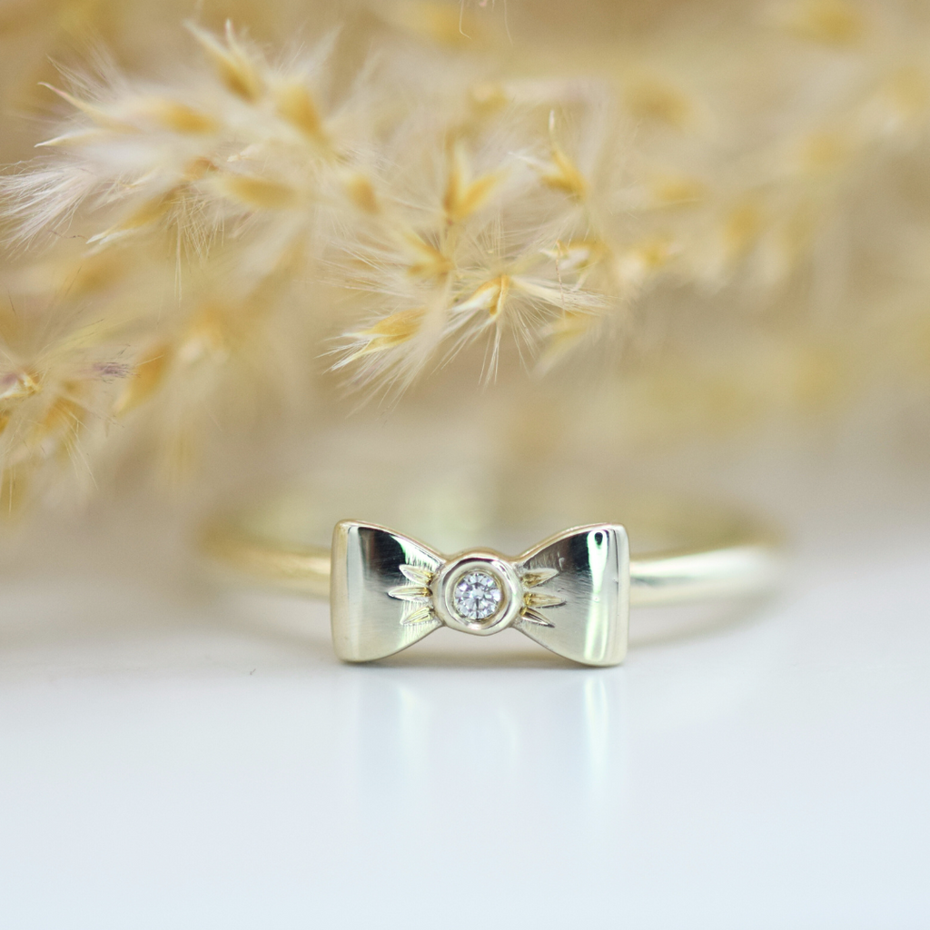 A 9k white gold ring with a central diamond and a bow-shaped design, placed on a white surface with dried flowers in the background.