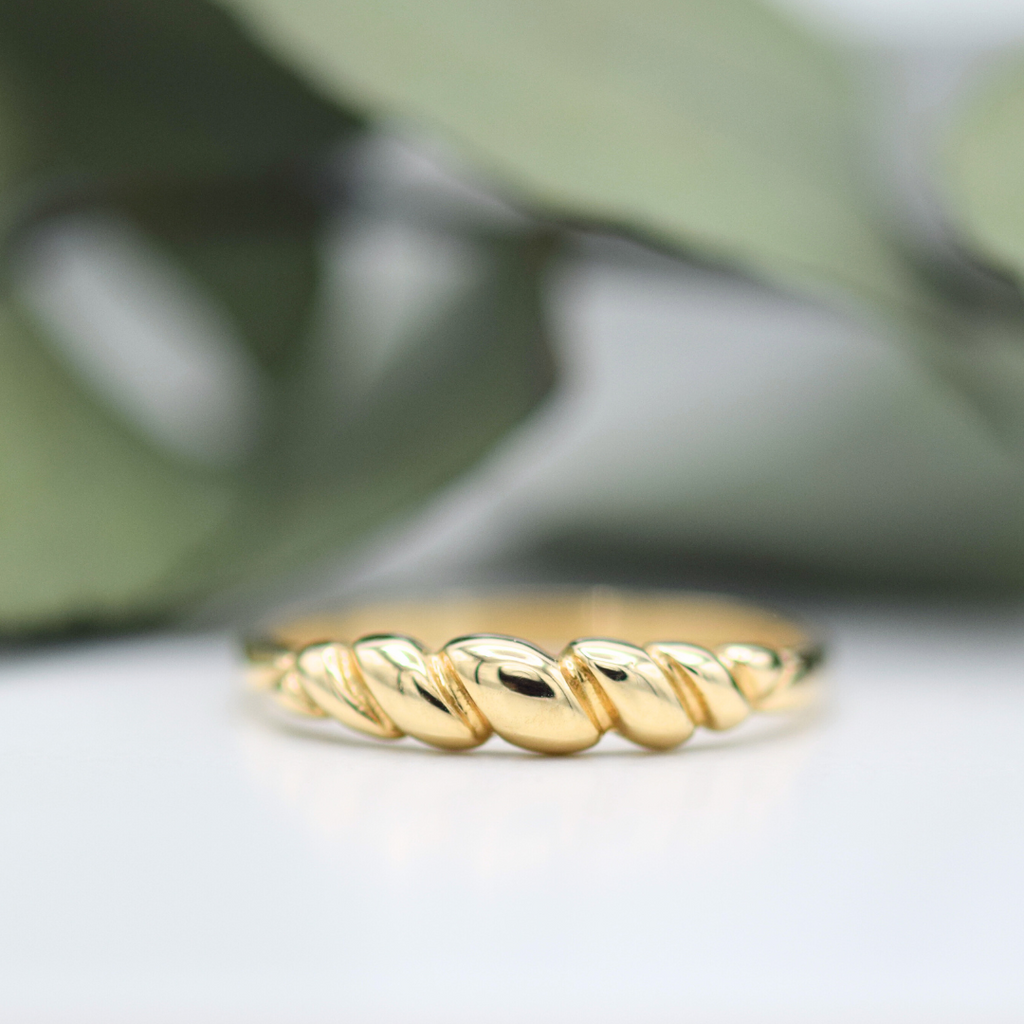 A gold twist ring on a white surface, with a blurred green leaf in the background.