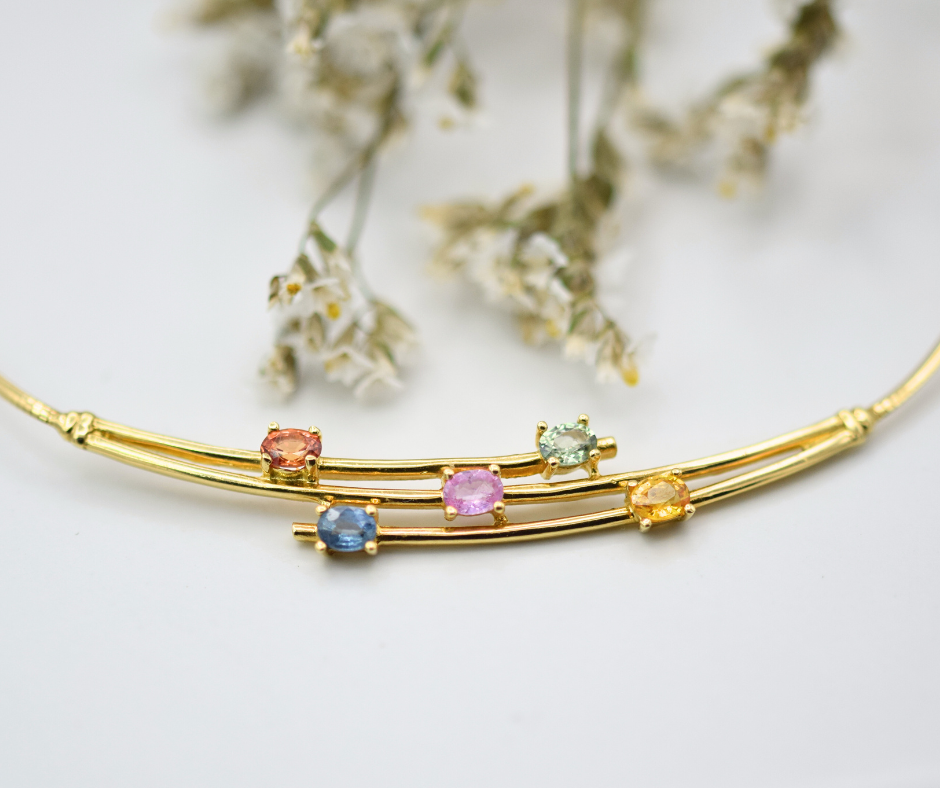 An 18k yellow gold necklace with five oval sapphires of mixed colors, displayed against a light background with some dried flowers.