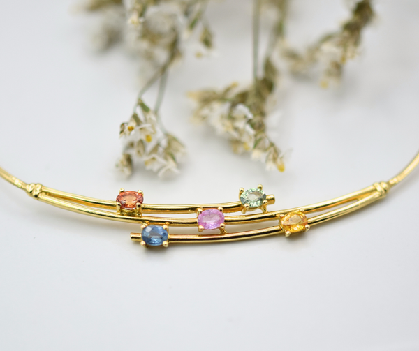 An 18k yellow gold necklace with five oval sapphires of mixed colors, displayed against a light background with some dried flowers.